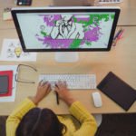 a person sitting at a desk with a computer and a screen with a drawing on it